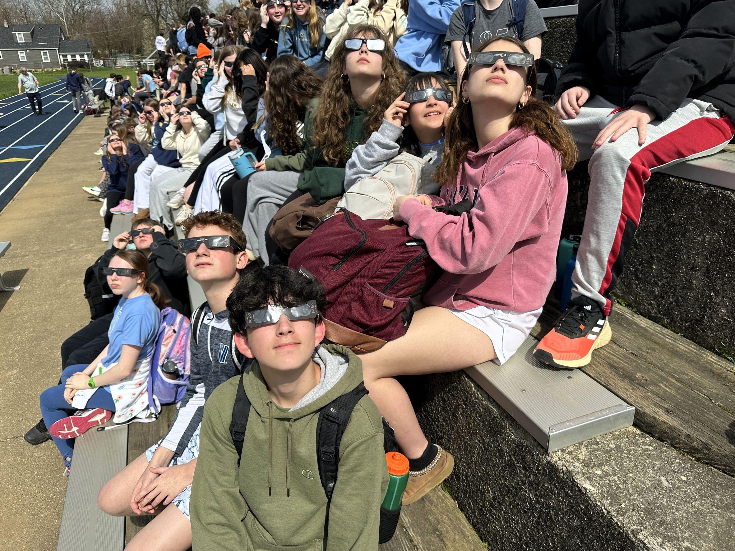 A large group of kids (over 50) sitting on the bleachers all wearing their eclipse glasses looking up at the sun